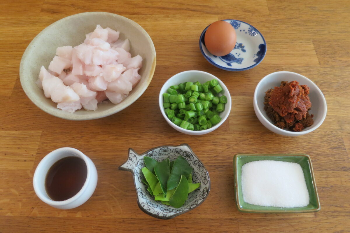 "Fresh ingredients for Thai fish cakes, including white fish fillets, red curry paste, kaffir lime leaves, green beans, egg, rice flour, and seasonings