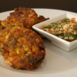 Plate of golden Thai corn fritters served with a small dish of cucumber relish on the side.